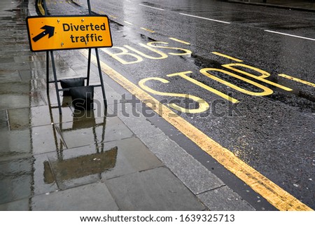 Diverted traffic black and yellow sign on wet pavement, next to road with Bus Stop text, lights reflect in rain covered asphalt