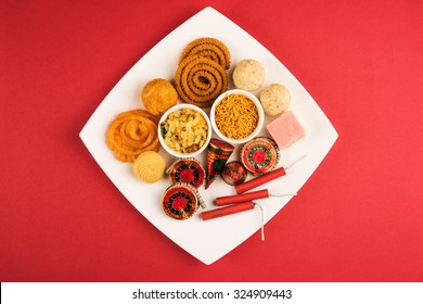 Diwali mithai like jalebi, bundi laddu, burfi and pera with Diwali snacks like chivda, sev, chakli served with Diwali firecrackers or fire crackers in a square white plate isolated on red background