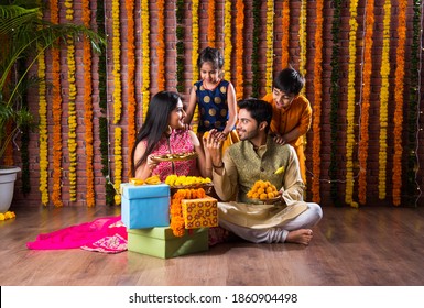 Diwali Or Rakshabandhan Celebration - Indian Young Family Of Four Celebrating Deepavali Or Bhai Dooj Festival With Sweet Laddoo, Oil Lamp Or Diya And Gift Boxes, Eating Food Or Taking Selfie