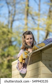 DIY Concept - Young Woman Holding A Hammer Working On A Wooden Playhouse Wearing Protective Gloves Outside In Backyard.