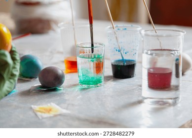 DIY Easter Egg Coloring Process. A brush for painting is washed in a glass with water. Nearby are several painted Easter eggs and glasses with paints