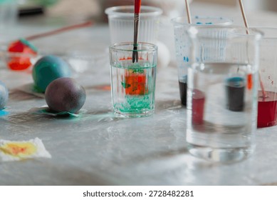 DIY Easter Egg Coloring Process. A brush for painting is washed in a glass with water. Nearby are several painted Easter eggs and glasses with paints
