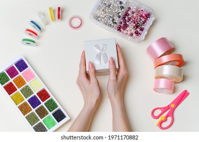 DIY Gift, Female Hands Are Holding A Gift Box. Crafts And Handicrafts.In The Background There Are Beads, An Embroidery Tool, Scissors, Ribbons. Do It Yourself. Holiday Gift