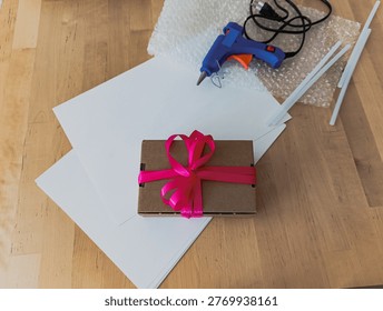 A DIY gift wrapping setup featuring a small cardboard box with a pink ribbon, a hot glue gun, glue sticks, and bubble wrap on a wooden table.