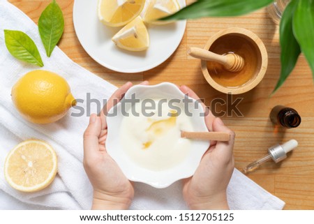 DIY natural face mask ingredients for bright and healthy skin. Woman hand holding a bowl of homemade face mask w/ yogurt, lemon, honey and essential oil on wood table. Beauty herbal skin care concept.