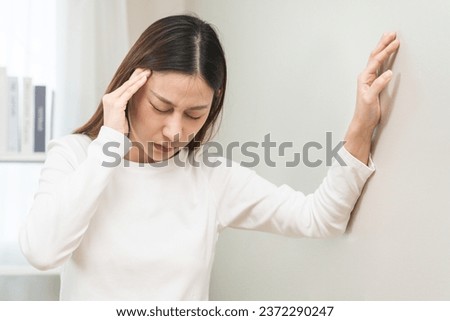 Dizzy asian young woman, girl headache or migraine pain. suffering from vertigo, difficulty standing up leaning on wall at home, holding head with hand, health problem of brain, inner ear not balance.