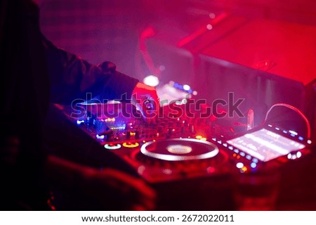 DJ adjusting mixer controls on an illuminated club deck bathed in red and blue stage lights.