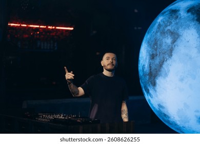 A DJ performs passionately in front of a luminous moon backdrop, creating an electrifying ambiance in a dimly lit venue. The mix of cosmic and artistic elements enhances the experience.