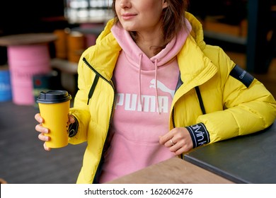 Dnipro, Ukraine - January 6, 2020: Young Woman Sitting In Cafe Hilding Cup Of Coffee To Go In Yellow Jacket Calvin Klein And Pink Hoodie Puma