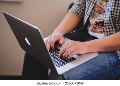 Dnipro, Ukraine - March 25, 2015: Close-up Of Office Worker In Plaid Shirt And Jeans, Typing On Apple MacBook Keyboard, Holding It On The Knees