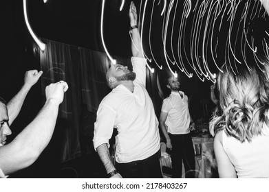 Dnipro, Ukraine - May 18, 2019. A Young Man Dances And Raises His Hand Up At A Party With Friends. Funny Emotions On A Holiday. Dancing And Bokeh From Lamps. Black And White Photography. Film Grain.