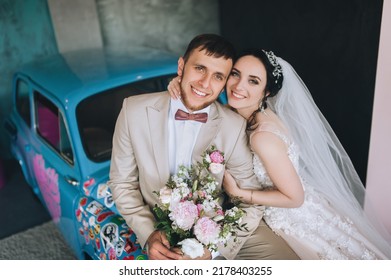 Dnipro, Ukraine - May 18, 2019.Smiling Groom Embraces A Beautiful Brunette Bride, Sitting On A Blue Vintage Car. Wedding Portrait Of Newlyweds In Love In A Modern Studio With A Beautiful Interior. 