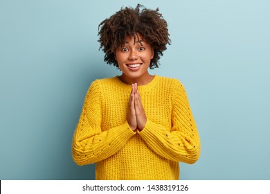 Do me favor. Cheerful Afro girl presses palms in pray gesture, asks for help, has gentle smile, grins and hopefully gazes at camera, beggs for advice, stands against blue background. Body language - Shutterstock ID 1438319126