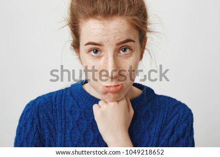 Do not be angry on me, honey. Gloomy beautiful girl with ginger hair and freckles looking from under forehead and sulking, leaning head on fist, whining or being upset, standing over gray wall