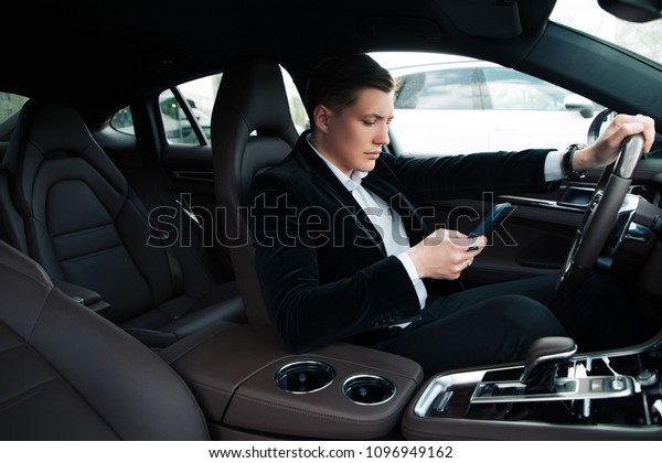 Do not type, while driving! Side view of the\
handsome smart young newcomer messaging, while using the phone and\
driving his automobile.