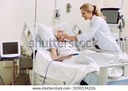 DO not worry. Friendly female medical worker smiling while helping her retired patient and adjusting an oxygen mask at hospital.
