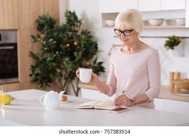 Do It With Pleasure. Joyful Nice Senior Woman Drinking Tea Ad Making Notes While Standing In The Kitchen