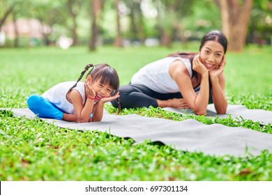 Do it together. Positive little cute girl sitting on the roll mat and practiving yoga with her mother while enjoying nice weather in the park - Powered by Shutterstock