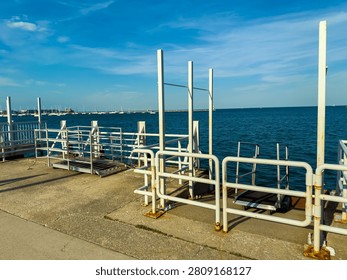 A dock area has empty platforms waiting for boats. The water is in front, and various boats are visible in the distance. The scene is bright and open.