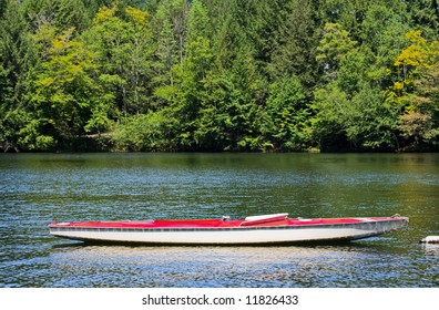 A Docked Sunfish Boat Without The Sail On A Lake. Green Trees Are In The Background.