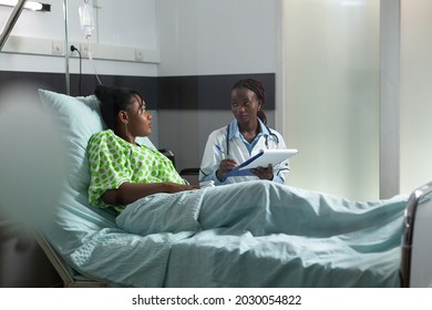 Doctor Of African Ethnicity Giving Disease Advice To Teenager Sitting In Hospital Ward Bed At Clinic. African American Medic Woman Helping Young Woman, Patient With Sickness Treatment