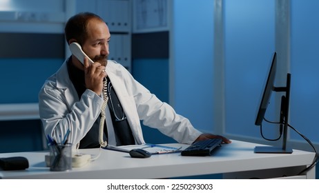 Doctor answering landline phone discussing sickness diagnosis with remote patient during checkup visit appointment. Physician working over hours at health care treatment to help cure disease - Powered by Shutterstock