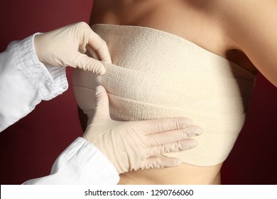 Doctor applying bandage on female chest after cosmetic surgery operation against color background