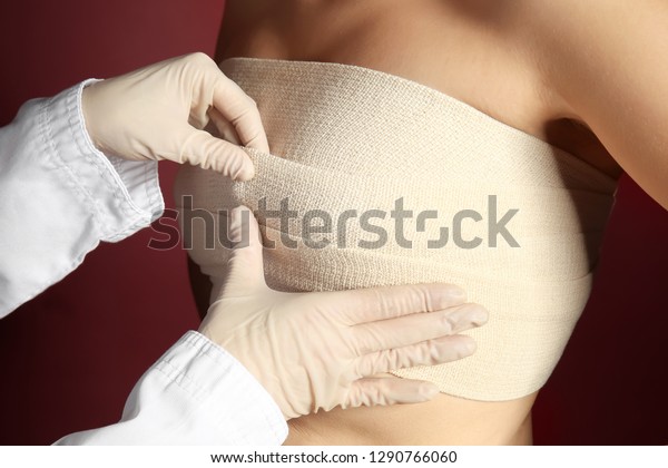 Doctor applying bandage on\
female chest after cosmetic surgery operation against color\
background