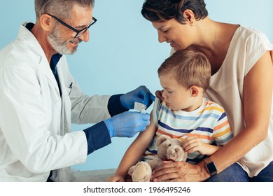 Doctor Applying Band-aid On Arm Of A Boy After Giving Him A Flu Shot. Boy Sitting With His Mother Getting Vaccination.