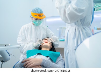 Doctor And Assistant In PPE Wear Protective Facemask Due To Covid19, Examining Young Woman Patient's Teeth Or Dental Checkup At Dental Clinic. Dentist Provide Oral Care Treatment And Medical Service.