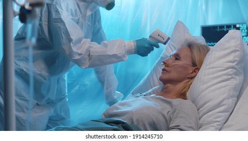 Doctor in biohazard suit measuring temperature of infected woman with infrared thermometer. Nurse in ppe overall checking patient temperature with scanner in hospital quarantine room