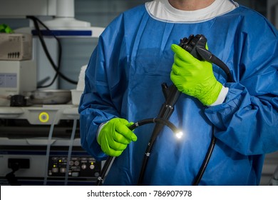 Doctor in blue protective clothing with green gloves is holding an endoscope in hand. The light on the endoscope is on, background with medicine utensils and instruments.