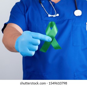 Doctor In Blue Uniform And Latex Gloves Holds A Green Ribbon As A Symbol Of Early Research And Disease Control, Symbol Of Lyme Disease, Kidney Transplantation And Organ Donation