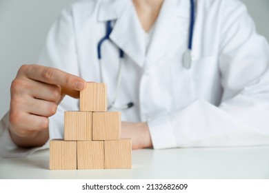 Doctor Building Pyramid Of Blank Wooden Cubes On White Table Against Light Background, Closeup. Space For Text