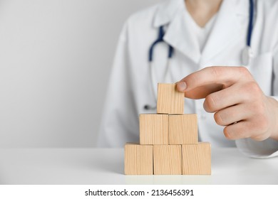 Doctor building pyramid of blank wooden cubes on white table against light background, closeup. Space for text
