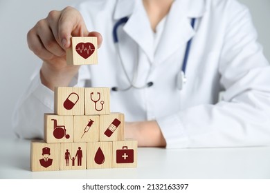 Doctor Building Pyramid Of Wooden Cubes With Different Icons At White Table, Closeup. Insurance Concept