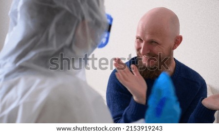The doctor came to the patient and amuses him in order to cheer up the patient. A doctor in a mask and protective suit is fooling around with a patient, a middle-aged man with a beard.