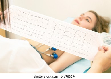 The Doctor Cardiologist Looks At The Printout Of The Cardiogram Of Patients, Which Lies On The Couch. Patient Girl On A Cardiogram Procedure.
