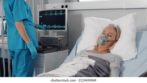 Doctor caring sick patient in hospital checking vital signs on monitor. Portrait of ill senior woman with oxygen mask resting in bed and nurse using computer on background checking physical condition