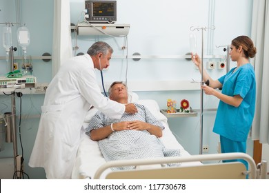 Doctor Checking Heart Beat Of Patient In Bed With Stethoscope With Nurse Adjusting I.V