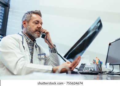 Doctor Checking A X-ray Report And Talking On Phone At Clinic. Radiologist Working At His Office Desk.