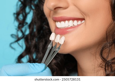 Doctor checking young woman's teeth color on light blue background, closeup. Dental veneers