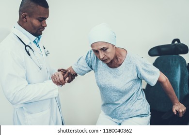 Doctor In Clinic.Doctor Helps Get Up. Patient Undergoes Rehabilitation. After Treatment For Cancer. Cancer Patient On A Wheelchair. Doctor With Stethoscope. Stethoscope On Neck. Scarf On Woman's Head.