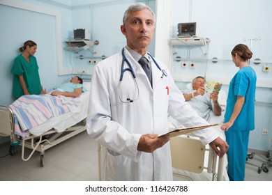 Doctor With Clipboard In Busy Hospital Room With Two Nurses Talking To Patients In Background, Healthcare Workers In The Coronavirus Covid19 Pandemic