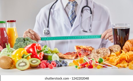 Doctor In Coat With Long Stethoscope Holding Ruler Over Table With Random Assortment Of Healthy And Unhealthy Food