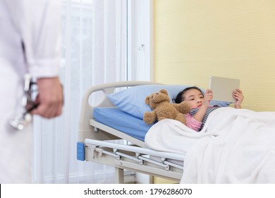 The Doctor Comes To Check Up A Little Girl Who Playing An IPad With A Bear On The Hospital Bed.