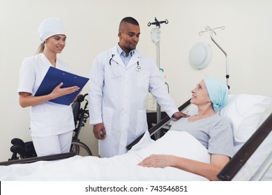 The doctor communicates with the patient. A woman is treated with cancer. Nearby is a nurse who holds a blank in her hand and records the patient's testimony. They are in a modern medical ward.
