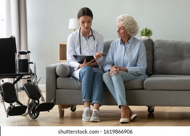 Doctor Consulting Disabled Older Woman, Filling Medical Form At Clipboard, Writing Prescription, Sitting On Couch At Home During Visit, Physician Wearing White Uniform Talking With Mature