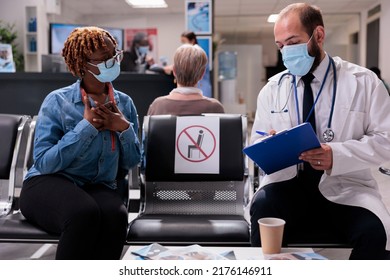 Doctor Consulting Female Patient In Medical Clinic Lobby, Sititng In Hospital Reception Waiting Area To Attend Checkup Appointment. Doing Health Examination During Covid 19 Pandemic.