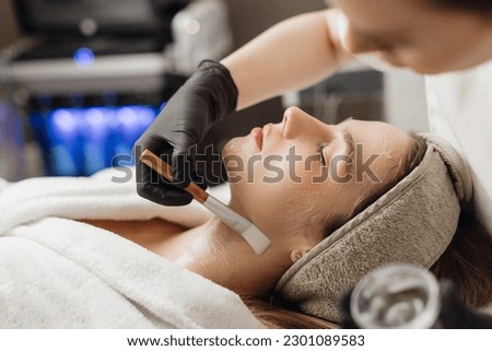 Doctor cosmetologist making face mask with brush in cosmetology clinic. Relaxed young woman getting professional facial treatment in spa salon